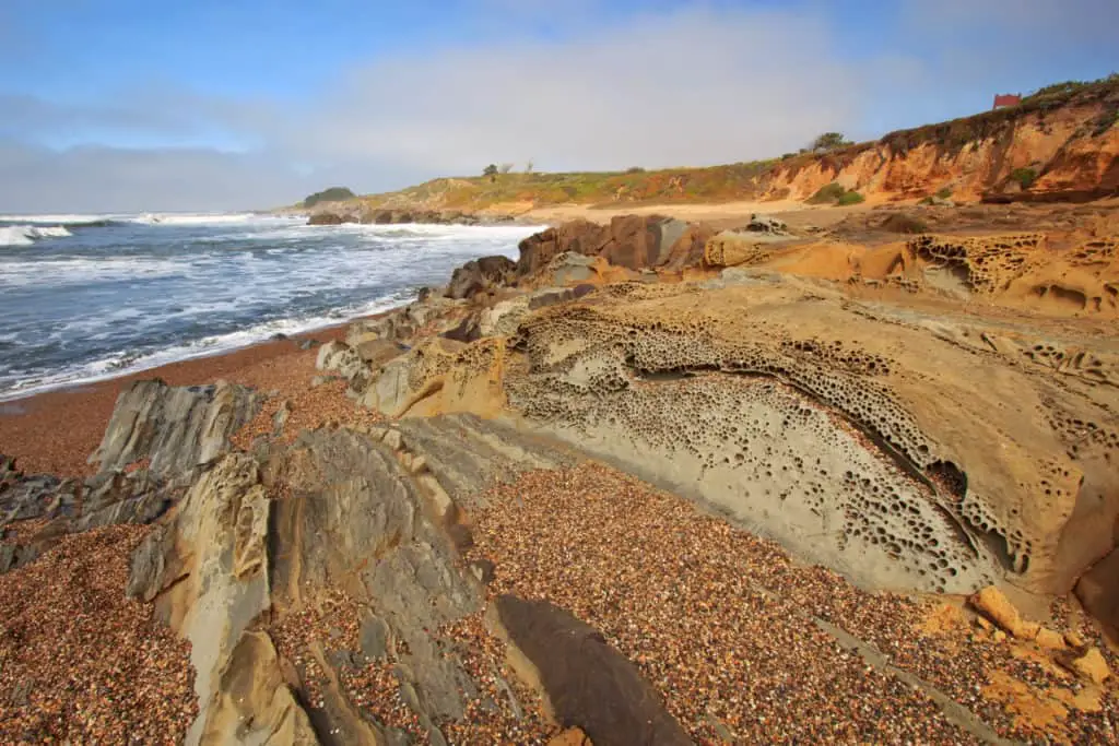 Pebble Beach and tafoni formations in Pigeon Point formation sandstone at Bean Hollow State Beach in San Mateo County California against a blue sky and white clouds - California Places, Travel, and News.