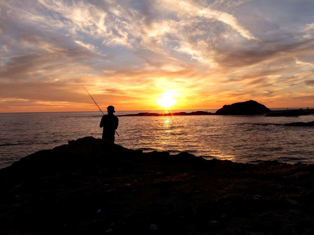 Silhouette of Fisherman holding a fishing pole at the beach at sunset in Southern California - California Places, Travel, and News.