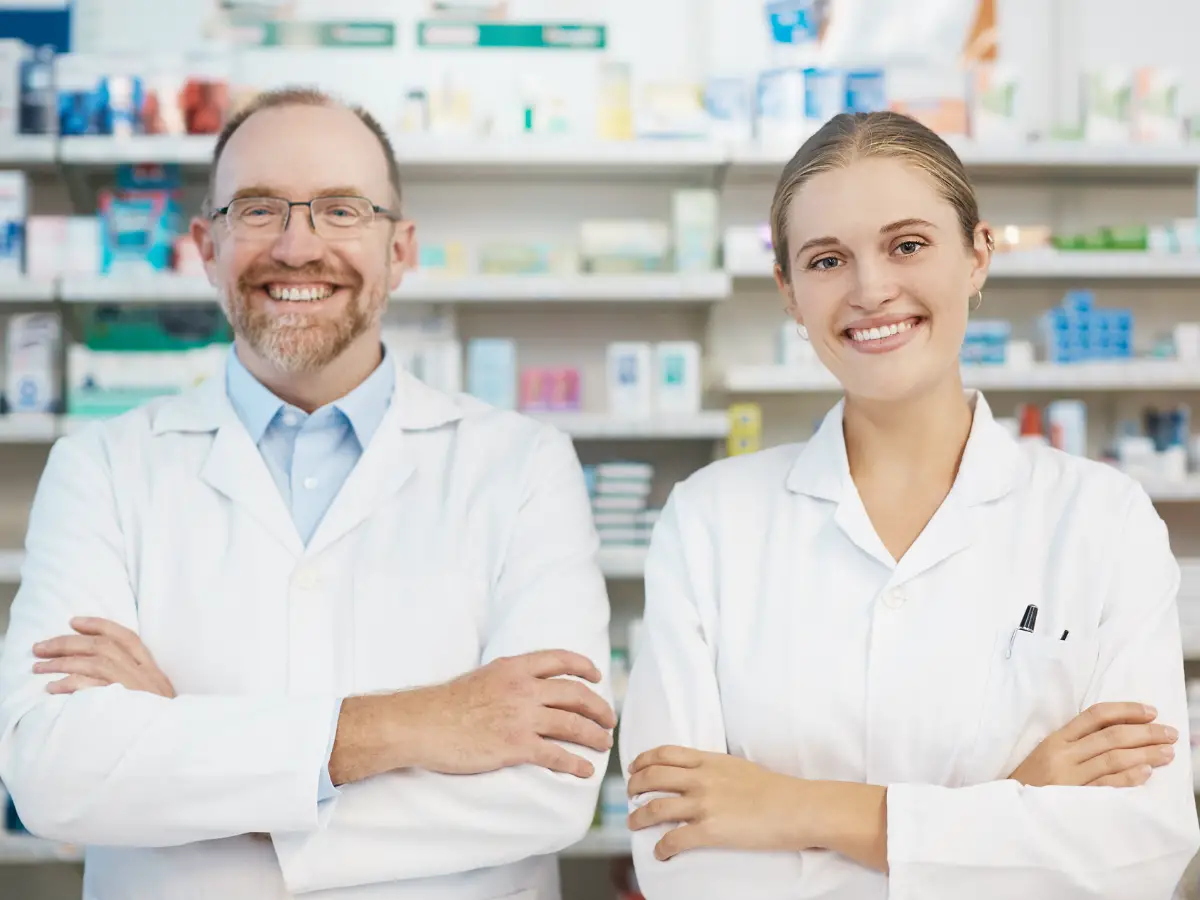 Senior and junior pharmacists stand behind dispensary counter smiling confidently 2 - California Places, Travel, and News.
